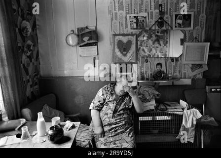 East End London 1970er Great Eastern Buildings Peabody Housing Estate. Eine Assistenzärztin Mrs. Edna Clarke in ihrem Wohnzimmer bedeckt ihre Augen, Kopf in Hand, erschöpft nach einem harten Arbeitstag. Sie trägt einen typischen 1970er-Jahre-Hausmantel, ein Overall, der ihre besten Kleidungsstücke bedeckt, die sie oft trägt, anstatt eine Schürze für die Hausarbeit. Gestern stand eine leere Glas-Pint-Milchflasche und heute frischer pinta auf dem überfüllten Tisch, während der Tee braut. An den Wänden ihres Zimmers hängen Fotos von der Hochzeit ihrer Tochter und eine große herzförmige Dekoration mit Spitze, die sie daran erinnert, dass sie immer geliebt wird. 70S GB HOMER SYKES Stockfoto