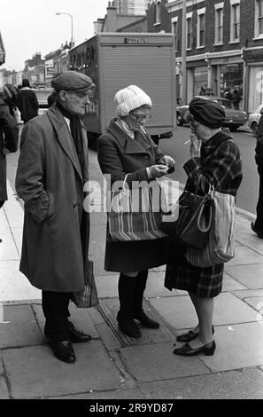 East End London 1970er Roman Road Market. Zwei Arbeiterinnen auf der Straße leuchten auf. Eine schlägt ein Streichholz aus einer Schachtel Streichhölzer für die anderen Zigaretten, während ihr Mann zusieht. Er trägt die Einkaufstasche und trägt eine flache Mütze und einen Schal, die auf traditionelle Weise getragen und gefesselt werden. Sie gehen einkaufen auf dem Roman Road Markt. Tower Hamlets, London, England um 1975. 70S GB HOMER SYKES Stockfoto