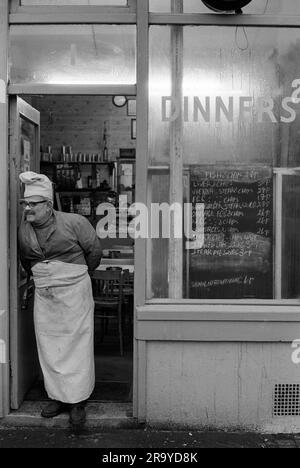 Brick Lane London 1970er. Der Besitzer mit Hut und Schürze des Küchenchefs, geknöpft an seiner Jacke, steht vor seinem schmierigen Löffel Dinner Café in Brick Lane und wartet auf die Mittagskunden. Die Speisekarte an der Tafel ist fast alles mit Pommes Frites. Whitechapel, London, England 1974. 70S GB HOMER SYKES Stockfoto