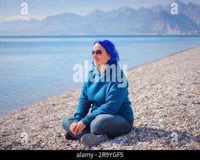 Eine Frau trägt einen hellblauen Hoodie und Jeans in Übergröße. Blauhaarige Frau draußen im Park am Strand von Antalya. Kleidungsmodell, leerer Platz für Text o des Stockfoto