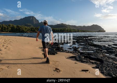 Gereifter Weißer Mann, Wanderer, nahe, Spaziergang entlang eines Sandstrands in der Nähe vulkanischer Lavafelsen im Ozean und einer Bergszene im Hintergrund Stockfoto