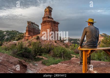 Ein erwachsener, weißer Wanderer sitzt auf einer Holzbank und beobachtet die herrlichen großen Felsentürme, den Leuchtturm, den Palo Duro Canyon State Park, TX Stockfoto