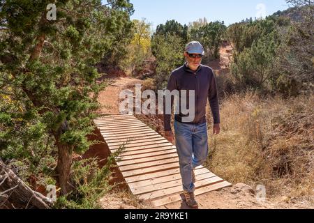 Ein erwachsener weißer Mann, der auf einer Holzbrücke über einen trockenen Bach spaziert und einen unbefestigten Pfad entlang führt, Kiowa Trail, Palo Duro State Park, Texas Stockfoto