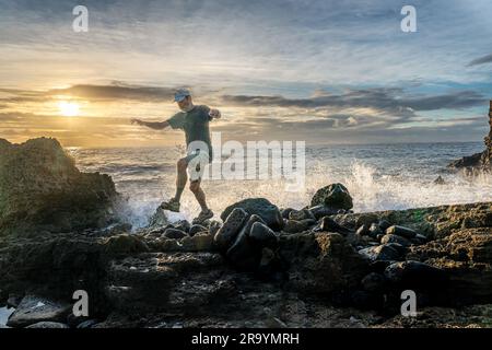 Ein Mann, der an einer Küste mit Basaltfelsen aus einem alten Lavafluss in die Brandung springt, während die Sonne hinter Wolken aufgeht, Poipu, Kauai, Hawaii Stockfoto