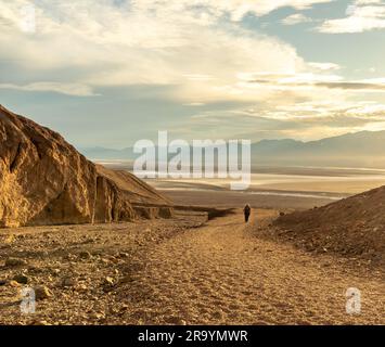 Ein Leihwanderer auf einem Weg in ein unteres Wüstental mit dramatischen Wolken, beleuchtet von der untergehenden Sonne, Natural Bridge, Death Valley National Park, Stockfoto