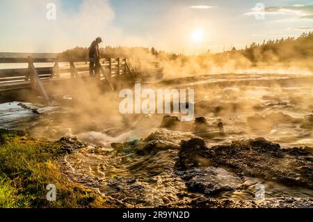 Ein Mann schaut auf eine wandelnde Holzbrücke an einem Thermalstrom nahe dem kochenden heißen Wasser, das die Sonne filtert, während sie im Hintergrund untergeht Stockfoto