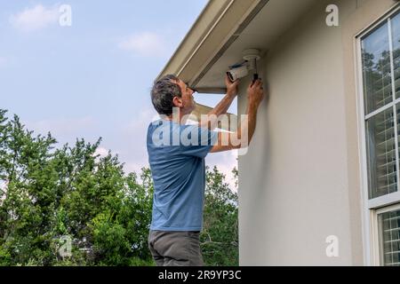 Weißer Weißer Mann, der eine Sicherheitskamera unter dem Haus repariert, mit Rinnen und Fenster sichtbar Stockfoto