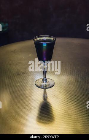 Gruseliges dunkelgraues Getränk in schillerndem Sherry-Glas auf Messingtisch bei Abendlicht Stockfoto