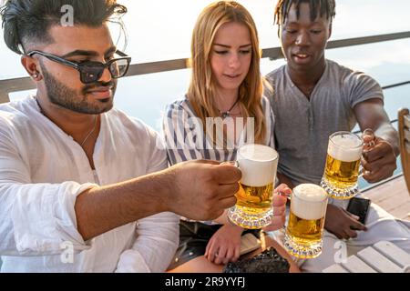 Kleine Gruppe von jungen erwachsenen multiethnischen Freunden, die mit drei Tassen Bier anstoßen - Sommerferienkonzept auf einem Schiff auf dem Fluss Stockfoto