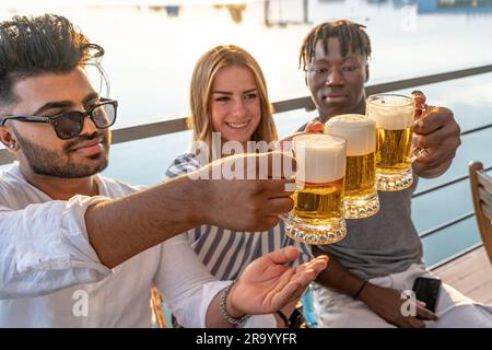 Kleine Gruppe von jungen erwachsenen multiethnischen Freunden, die mit drei Tassen Bier anstoßen - Sommerferienkonzept auf einem Schiff auf dem Fluss Stockfoto