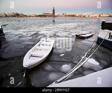 Festgefahrenes Boot im gefrorenen Wasser am Södermälarstrand mit Rathaus im Hintergrund. Stockholm, Schweden. Stockfoto