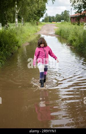 Ein junges Mädchen mit Gummistiefeln, das nach einer Überschwemmung in einer Wasserpfütze auf einer Landstraße spaziert. Stockfoto
