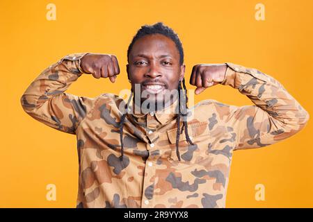 Ein afroamerikanischer Mann, der Trizeps vor dem Training im Studio auf gelbem Hintergrund nachgibt. Selbstbewusster Sportler, der sich posiert, um Bizeps zu zeigen, Muskelkörper von der Fitness Stockfoto