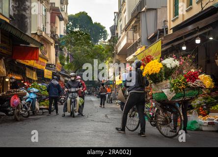 Eine Blumenverkäuferin schiebt ihr beladenes Fahrrad an Geschäften in Cau Go vorbei, innerhalb der Altstadt von Hanoi, Vietnam. Stockfoto