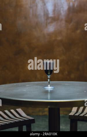 Schwarzes Holzkohle-Limonade-Getränk in schillerndem Sherry-Glas auf goldenem Messingtisch, Sommerabend im Freien Stockfoto