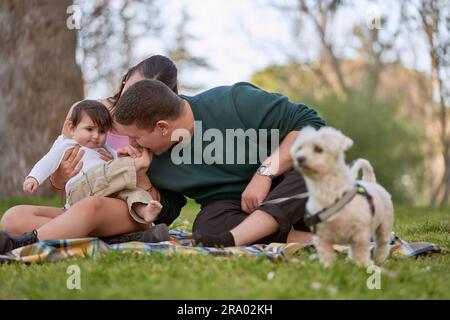 Ein junges Paar, das ein Picknick macht, auf dem Gras sitzt und die Füße seines Babys kitzelt Stockfoto