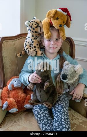 Porträt eines 7-jährigen Mädchens, das in einem Sessel sitzt, umgeben von Plüschtieren, einschließlich eines Hundes auf dem Kopf. HERR Stockfoto