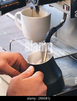 Ein professioneller Barista gießt aufgeschäumte Milch aus einer Kaffeemaschine in eine Kanne Stockfoto