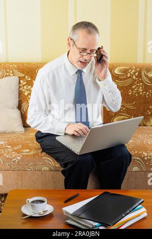 Ein gutaussehender Geschäftsmann, der zu Hause mit einem Computer arbeitet, mit seinem Handy einen Partner anruft und bereit ist, eine heiße Tasse schwarzen Kaffee zu trinken Stockfoto