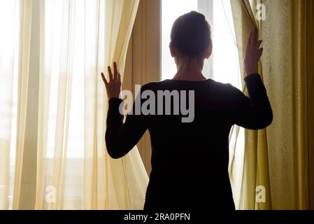 Eine Frau in schwarz zieht den Vorhang beiseite nach Trog die Fenster schauen Stockfoto