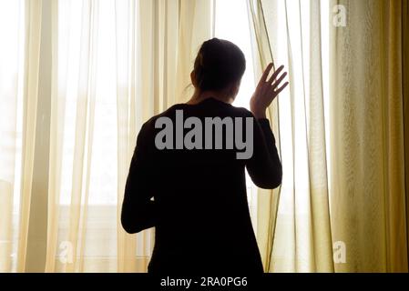 Eine Frau in schwarz zieht den Vorhang beiseite nach Trog die Fenster schauen Stockfoto