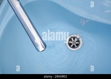 Das Wasser fließt aus dem Hahn in einem blauen Waschbecken. Die Flüssigkeit gießt, wirbelnden, in den offenen Stecker Stockfoto