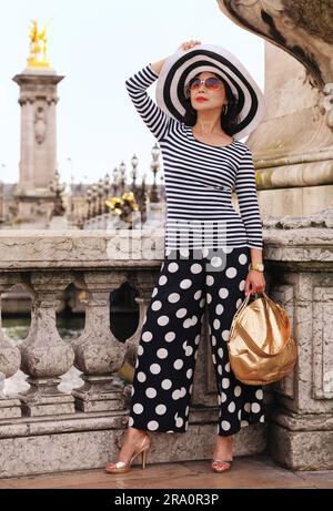 Eine glamouröse, reife Frau auf der Aussichtsplattform mit Blick auf die kunstvoll verzierten Jugendstillampen und Skulpturen von Pont Alexandre III trägt Streifen und Punkte Stockfoto