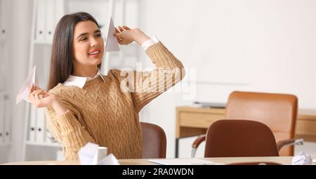 Junge Frau, die Papierflugzeuge an den Tisch im Büro wirft Stockfoto