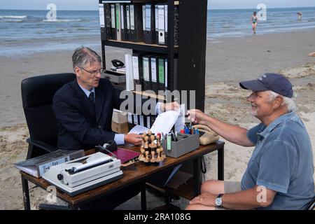Spiekeroog, Deutschland. 29. Juni 2023. Albrecht Fersch (l) sitzt an seinem Schreibtisch am Strand der ostfriesischen Insel Spiekeroog und spricht mit dem Touristen Falko Grunau. Mit seinem Projekt "Schreibtischtäter" möchte der Actionkünstler Humor einsetzen, um Menschen dazu zu ermutigen, über soziale Bedingungen und die menschliche Existenz nachzudenken. Kredit: Marco Rauch/dpa/Alamy Live News Stockfoto