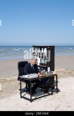 Spiekeroog, Deutschland. 29. Juni 2023. Albrecht Fersch sitzt an seinem Schreibtisch am Strand der ostfriesischen Insel Spiekeroog. Mit seinem Projekt "Schreibtischtäter" möchte der Actionkünstler Humor einsetzen, um Menschen dazu zu ermutigen, über soziale Bedingungen und die menschliche Existenz nachzudenken. Kredit: Marco Rauch/dpa/Alamy Live News Stockfoto