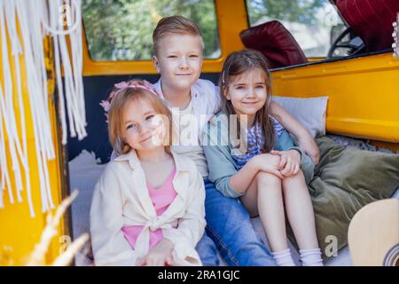 Drei süße kleine Kinder sitzen im Retro-Minivan im Bo-Stil. Kinder spielen und haben Spaß auf weichen Pilows im Auto. Fröhliche Schwestern und Braut Stockfoto