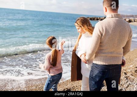 Glückliche junge Familie am Meer. Ein Mann und eine Frau halten Händchen und sehen ihre Tochter an. Ein kleines Mädchen in Pullover und Jeans wirft Kiesel in die Luft Stockfoto