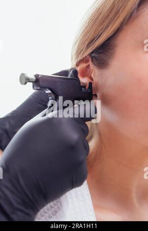 Nahaufnahme des Gesichts einer jungen blonden Frau, die eine Ohrpiercing-Operation durchführt. Arzthandschuhe in schwarzen Handschuhen halten eine Ohrstechpistole. Medizinisches Personal macht Loch an Stockfoto