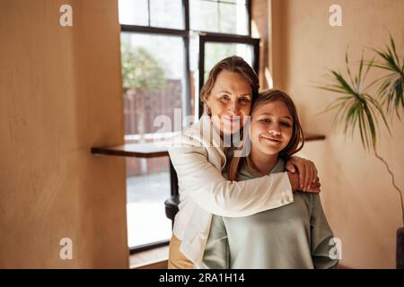Eine schöne Frau mittleren Alters in einer weißen Jacke lächelt und umarmt ihre Enkelin. Ein Mädchen im Teenageralter in lässigen Klamotten posiert mit ihrer Großmutter in einem Zimmer Stockfoto