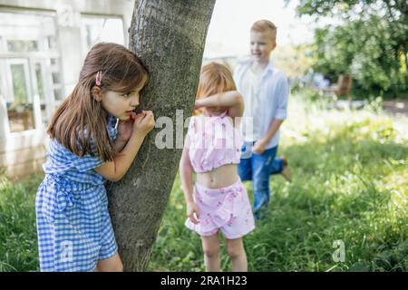 Kleine Kinder können sich im Garten verstecken. Süßes Mädchen versteckt sich hinter dem Baum. Der Junge sucht ein Versteck. Das hübsche Kind hat ihr Ei geschlossen Stockfoto