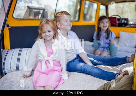 Drei süße kleine Kinder sitzen im Retro-Minivan. Kinder lächeln und haben Spaß auf der weichen Matratze im Auto. Fröhliche Schwestern und Bruder warten Stockfoto