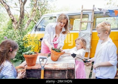 Eine junge Frau und ihre drei Kinder Pflanzen Blumen in Töpfen im Garten. Hübsche Mutter, süße Mädchen und Junge sind im Gartenbau beschäftigt. Glückliche Familie Stockfoto