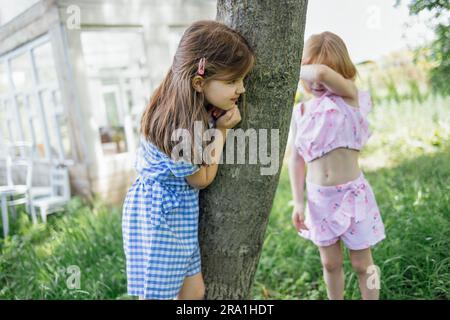 Kleine Kinder können sich im Garten verstecken. Ein süßes Mädchen versteckt sich hinter einem Baum. Ein hübsches Kind hat die Augen geschlossen und wartet auf sie Stockfoto