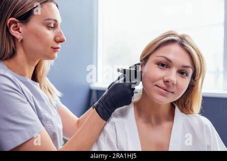 Nahaufnahme des Gesichts einer jungen blonden Frau, die eine Ohrpiercing-Operation durchführt. Arzthandschuhe in schwarzen Handschuhen halten eine Ohrstechpistole. Medizinisches Personal macht Loch an Stockfoto