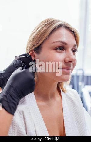 Nahaufnahme des Gesichts einer jungen blonden Frau, die eine Ohrpiercing-Operation durchführt. Arzthandschuhe in schwarzen Handschuhen halten eine Ohrstechpistole. Medizinisches Personal macht Loch an Stockfoto