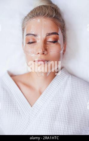 Frau mit transparenter Vitaminmaske im Gesicht im Spa-Salon oder in der Klinik. Blondes, hübsches Mädchen im weißen Bademantel wird einer feuchtigkeitsspendenden Behandlung unterzogen Stockfoto