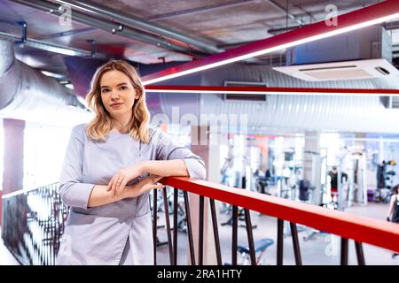 Junge attraktive Frau in grauer Uniform steht vor dem Hintergrund des Fitnessstudios. Eine hübsche lächelnde blonde Masseuse arbeitet in einem Fitnessclub. Sport-Equi Stockfoto