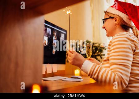 Die junge Frau mit weihnachtsmann-Hut kommuniziert gerne per Videoverbindung mit Freunden. Ein lächelndes Mädchen hält ein Glas Champagner und Glitzer in den Händen und gratuliert Stockfoto