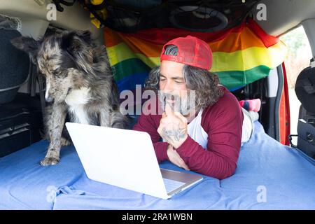 Ein Mann mittleren Alters, der mit einem Laptop im Wohnwagen liegt. Reisender mit Remote-Computer, der online arbeitet und Eitelkeit und Freiheit genießt, ruht sich im Outdoor-Camp aus Stockfoto