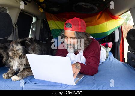 Ein Mann mittleren Alters, der mit seinem Haustier im Campingwagen liegt und einen Laptop benutzt. Ein Reisender mit einem Remote-Computer, der online arbeitet und Spaß hat Stockfoto