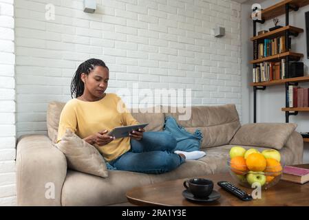 Junge afroamerikanische Frau sitzt auf der Couch im Wohnzimmer und surft online auf einem Tablet, fröhliche, birassistische Frauen ruhen sich zu Hause auf dem Sofa aus Stockfoto