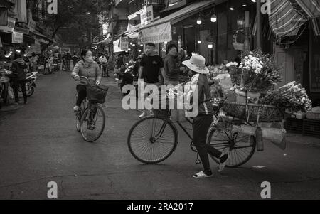 Eine Blumenverkäuferin schiebt ihr beladenes Fahrrad an einem anderen Radfahrer und Geschäften in Cau Go vorbei, innerhalb der Altstadt von Hanoi, Vietnam. Stockfoto