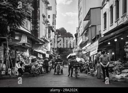 Eine Blumenverkäuferin schubst ihr beladenes Fahrrad vorbei an geschäftigen Geschäften in Cau Go, in der Altstadt von Hanoi, Vietnam. Stockfoto