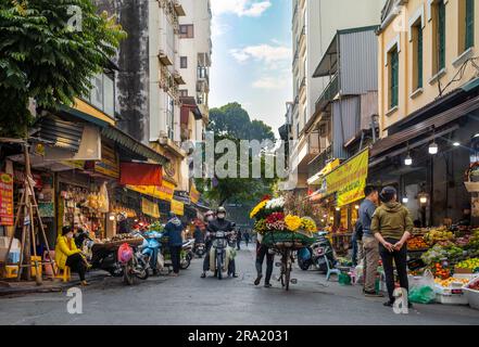 Eine Blumenverkäuferin schubst ihr beladenes Fahrrad vorbei an geschäftigen Geschäften in Cau Go, in der Altstadt von Hanoi, Vietnam. Stockfoto