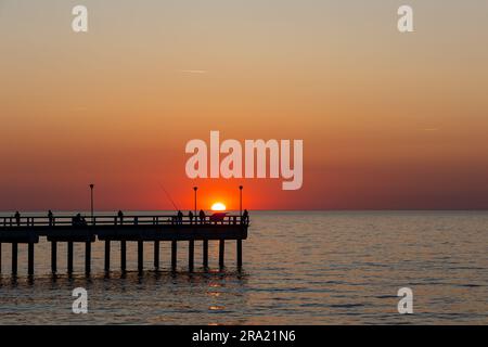 Ein großer orangefarbener Sonnenuntergang über einem ruhigen Meer. Fischer, die am Pier angeln. Statische Aufnahme während eines wolkenlosen Abends Stockfoto